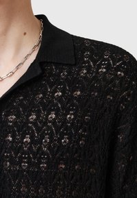 Camisa de encaje negro con un patrón bordado, cuello tipo solapa y una textura delicada. Accentuada con un collar de cadena plateada.