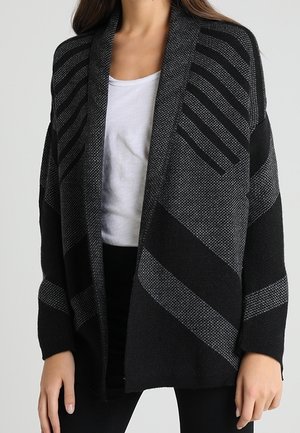 Femme portant un cardigan noir et gris ouvert à l'avant avec des motifs diagonaux et rayés, sur une chemise blanche et un pantalon noir.