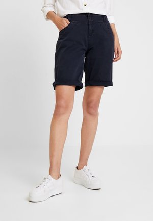Person iført opkørte mørkeblå shorts, hvid skjorte med knapper og hvide plateau-sneakers, stående med den ene hånd i lommen.