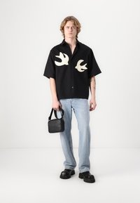 Camisa preta de manga curta com desenhos de andorinhas brancas, combinada com jeans azul claro e sapatos pretos chunky, segurando uma pequena bolsa preta.