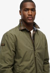 Olive green bomber jacket with zip closure, featuring a plaid lined collar in navy, orange, and green. Includes logo patch on sleeve.