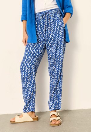 Woman wearing blue and white wavy patterned pants, blue shirt, white top, and beige double-buckle sandals standing with hands in pockets.