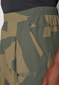 Olive green shorts with a camo pattern, elastic waistband, side pocket, and embroidered "TERREX" logo in grey. Lightweight fabric texture.