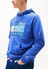 Blauer Baumwoll-Hoodie mit einem Frontgrafik, die den Text "TOMMY JEANS" zeigt. Er hat eine Kordelzugkapuze und eine Kängurutasche; gerippte Bündchen und Saum.