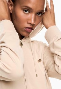 Joven con una sudadera con cremallera beige, con las manos cerca de la cabeza, mirando directamente hacia adelante con expresión neutral, contra un fondo blanco.