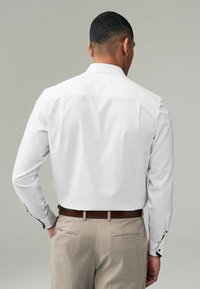 Weißes Langarmhemd mit glatter Textur; brauer Gürtel; beige Hose mit detaillierten Manschetten und Knopfakzenten.