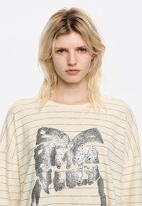 Maglietta leggera beige, a maniche lunghe, con sottili righe blu navy e una stampa grafica metallica del testo "BIMBA Y LOLA" sulla parte anteriore. Vestibilità morbida e rilassata.