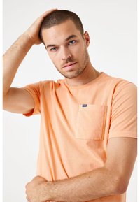 Camiseta de algodón naranja claro con mangas cortas y un bolsillo en el pecho. Presenta un cuello redondo y una textura suave.