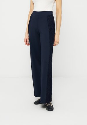 Femme portant un pantalon large taille haute bleu marine avec des panneaux latéraux en dentelle, un haut blanc sans manches et des mocassins noirs avec un détail doré.