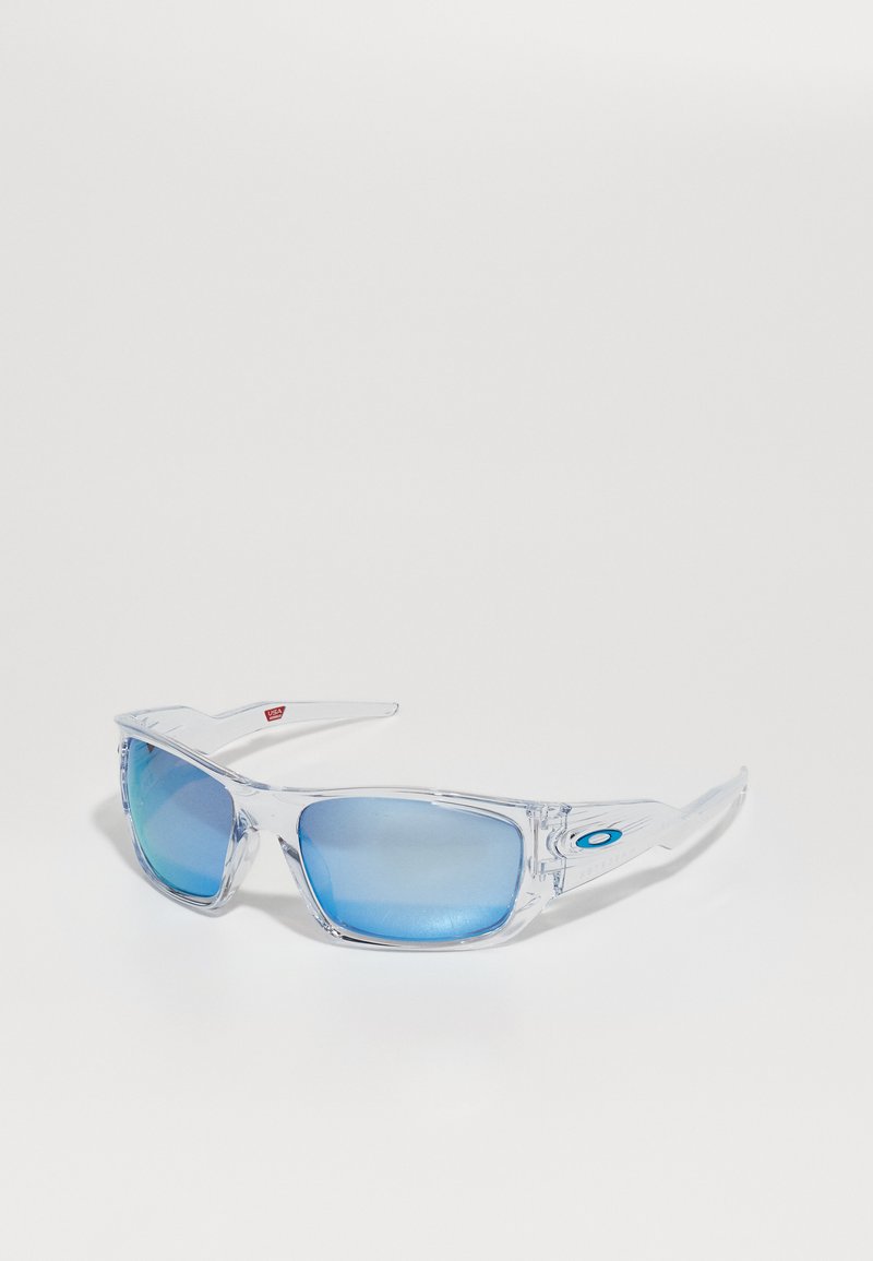Occhiali da sole con montatura trasparente, lenti blu specchiate, design angolare e accento del logo blu sul lato, caratterizzati da una forma aerodinamica.