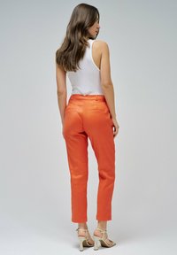 Mujer con cabello largo castaño, vistiendo una blusa sin mangas blanca, pantalones naranjas y sandalias de tacón beige, de pie y de espaldas.