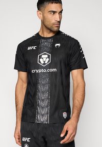 Μαύρο μπλουζάκι UFC με υφασμένη υφή, στρογγυλή λαιμόκοψη και κοντά μανίκια. Διαθέτει το λογότυπο και τη σήμανση του crypto.com στο αριστερό στήθος και στα μανίκια.