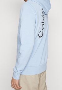 Felpa leggermente blu con cappuccio e coulisse, con un grande logo nero "Calvin" sulla schiena, realizzata in tessuto morbido con orlo a coste.