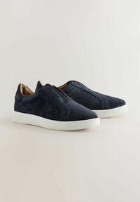 Zapatos de ante azul marino tipo slip-on con suelas blancas, que presentan un detalle de costura a lo largo de los laterales, sobre un fondo claro.