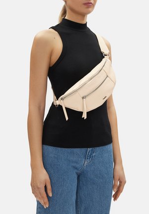 Žena v bezrukávovém černém topu a modrých džínách s béžovou kabelkou přes rameno, která má dvě přihrádky na zip.