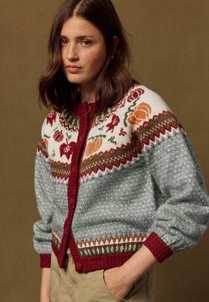 Junge Frau, die eine gemusterte Wollstrickjacke mit floralen und geometrischen Designs in Rot-, Orange-, Grün- und Grautönen trägt, steht mit einer Hand in der Tasche.