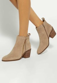Beige suede ankle boots with a rounded toe, a side zipper, and a stacked heel. Features a pull tab for easy wear.