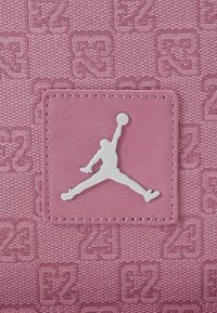 Το ροζ υλικό με υφή διαθέτει ανάγλυφο σχέδιο. Ένα τετράγωνο δερμάτινο ταμπελάκι παίζει το λογότυπο Jumpman σε λευκό, τονίζοντας την ταυτότητα της μάρκας.