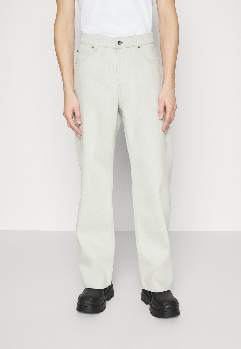 Pantalones de denim gris claro con un ajuste holgado, diseño de cinco bolsillos, cierre frontal con botón y tejido texturizado, acompañados de zapatos negros gruesos.