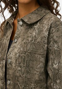 Veste imprimée serpent marron avec un col, des boutons-pression et deux poches avant. Le tissu a une apparence texturée et douce.