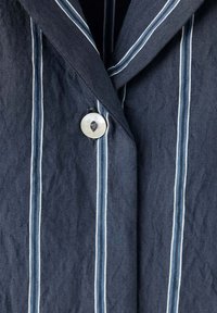 Gros plan d’un tissu bleu foncé avec des rayures verticales blanches et bleu clair et un seul bouton blanc cousu près d’un col.