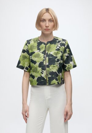 Femme aux cheveux blonds courts portant un haut zippé vert et noir à manches courtes avec motif floral et un pantalon blanc taille haute sur fond blanc.