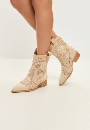 Beige gewebte Stiefeletten mit floralen Häkelmustern und niedrigen Holzabsätzen, getragen an überschlagenen Beinen vor einfarbigem Hintergrund.