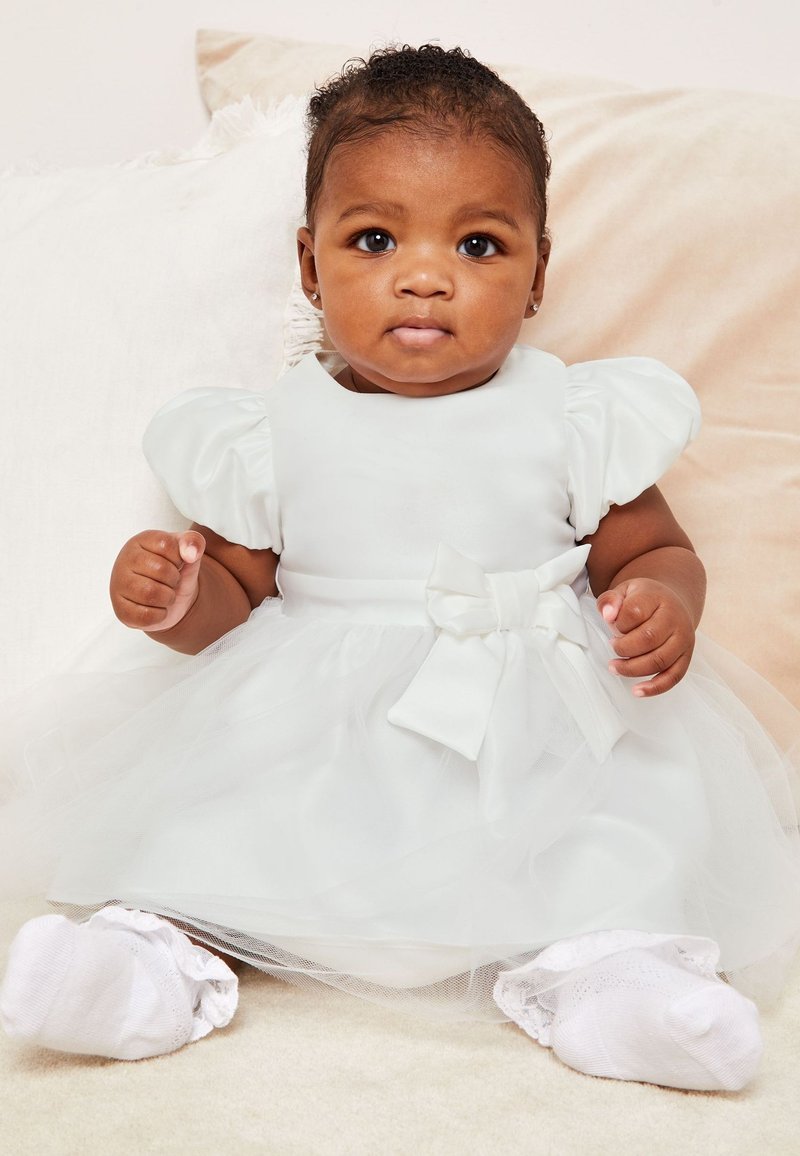 Bambino che indossa un vestito bianco con maniche a sbuffo e fiocco, seduto su una coperta chiara, guarda dritto davanti a sé.