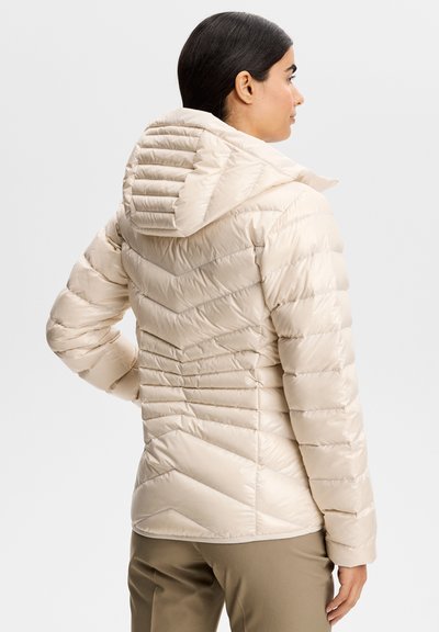 Femme portant une veste matelassée beige clair avec capuche, vue de dos, debout avec une main sur la hanche.