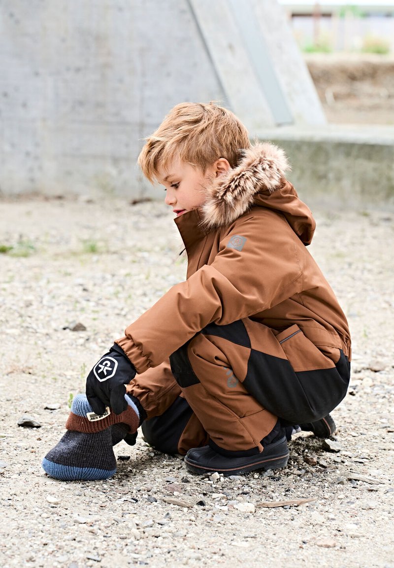 Bambino con giacca invernale marrone con cappuccio di pelliccia si accovaccia all'aperto, indossando un calzino lavorato a maglia scuro sopra una scarpa su terreno ghiaioso.