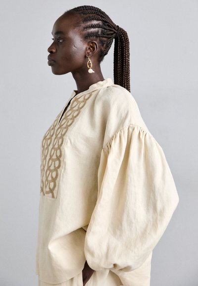 Femme avec des cheveux tressés en queue de cheval, portant un chemisier crème à manches bouffantes et broderies décoratives, se tenant de profil contre un fond uni.