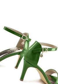 Grüne Lackleder-Pumps mit einem Knöchelriemen, glänzender Oberfläche, schmalem hohen Absatz und einem kleinen Markenanhänger.