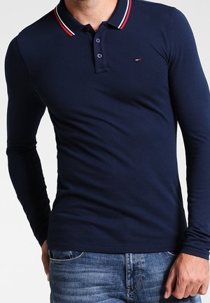 Homme portant un polo bleu marine ajusté à manches longues avec un col rayé rouge et blanc et un jean bleu, debout avec une main dans la poche.