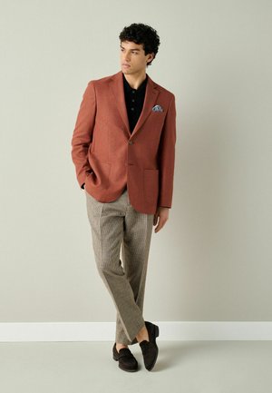 Man wearing rust-colored blazer, black shirt, checkered beige pants, and dark loafers, standing with hands in pockets against plain wall.