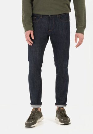 Dark blue denim jeans with a slim fit, featuring orange stitching and rolled cuffs, paired with olive green sneakers and a matching sweater.