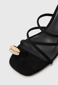 Sandalia de tacón de ante negro con tiras entrelazadas, punta cuadrada y herrajes de acento en oro y plata en la tira del dedo. Suela interior suave.