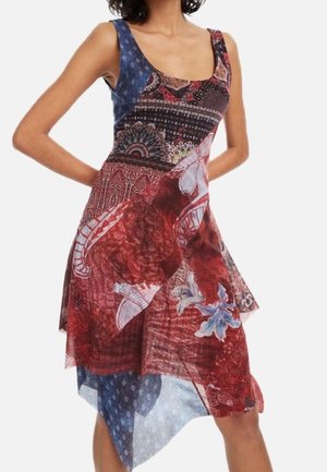 Femme portant une robe sans manches avec des motifs floraux et géométriques mélangés en rouge, bleu et noir, avec un ourlet asymétrique.