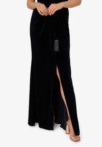 Długa, granatowa sukienka z aksamitnego materiału z klinem na przodzie, ozdobiona srebrnym frędzlem. Połączona ze złotymi sandałami na wysokim obcasie z paskami.