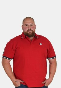 Polo rouge avec un col, arborant un logo sur la poitrine et des accents bleu marine sur les manches, porté avec un jean bleu.