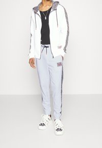 Weißer Reißverschlussjacke mit schwarz gemusterten Ärmeln, schwarzem Tanktop, hellgrauen Jogginghosen mit passenden Akzenten und weißen Sportschuhen.