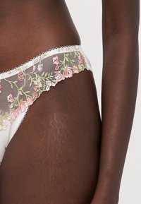 Weißer Spitzenstring mit floralem Motiv in Rosatönen und Grüntönen, ausgestattet mit einem durchsichtigen Netzpaneel und gewellten Kanten. Weicher Stoff.