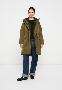 Parka verde oliva con un interno morbido e texturizzato, cappuccio regolabile e tasche frontali. Abbinato a jeans blu con risvolto e scarpe nere.