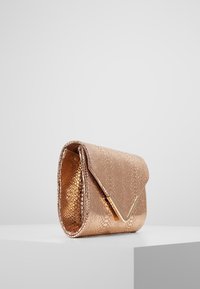 Clutch kabelka v odstínu růžového zlata s texturovaným povrchem a hladkým dokončením, která má na předním klopě V-tvarovaný zlatý kovový detail. Kompaktní a stylový design.