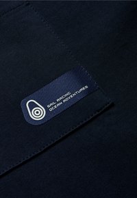Tela azul marina con una etiqueta texturizada que presenta un logo, "Sail Racing Ocean Adventures," en acentos de color blanco y azul claro.