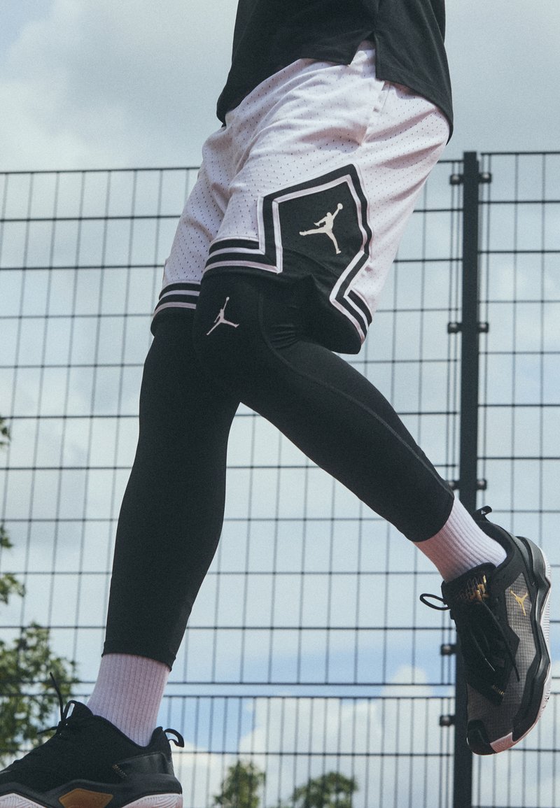 Shorts de basket noir à thème Jordan avec des accents blancs, leggings de compression noirs et baskets noires avec des détails dorés, en mouvement.