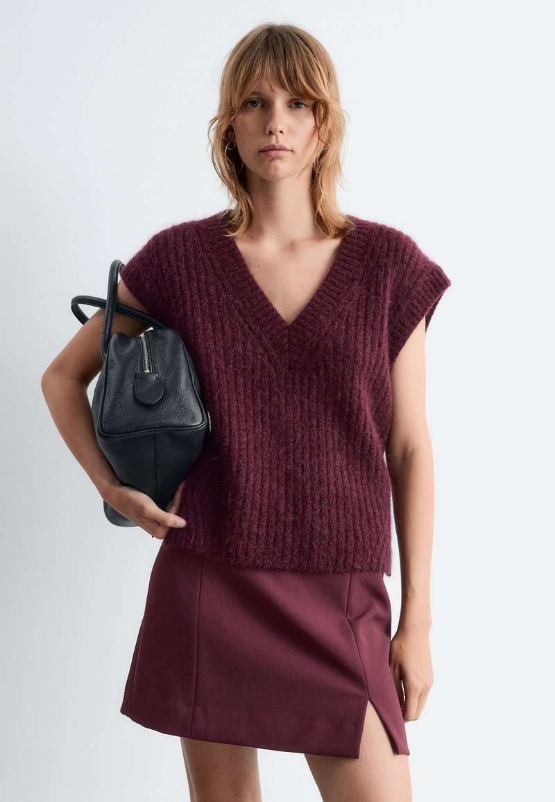 Burgunderfarbener, gestrickter Kurzarm-Pullover mit V-Ausschnitt, kombiniert mit einem burgunderfarbenen Minirock mit seitlichem Schlitz und einer schwarzen Lederhandtasche.