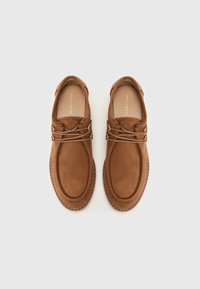 Chaussures en daim marron avec un bout arrondi, dotées de lacets maintenus par une quincaillerie en ton or, et d'une semelle en caoutchouc texturée. Design plat, sans accents de style visibles.
