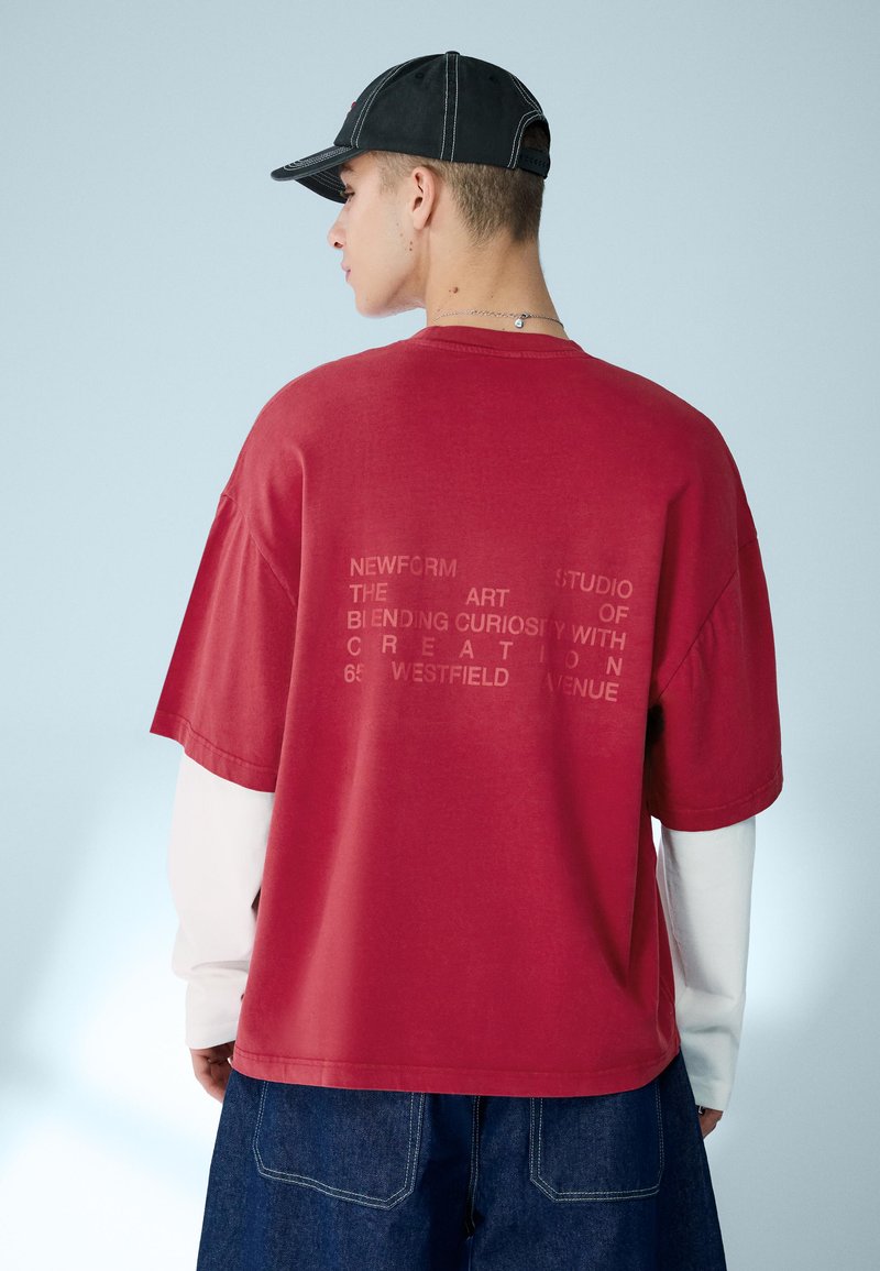 Mladý muž otočený zády, nosí červené oversized tričko s vybledlým nápisem, pod ním bílé dlouhé rukávy, tmavé džíny a černou čepici.