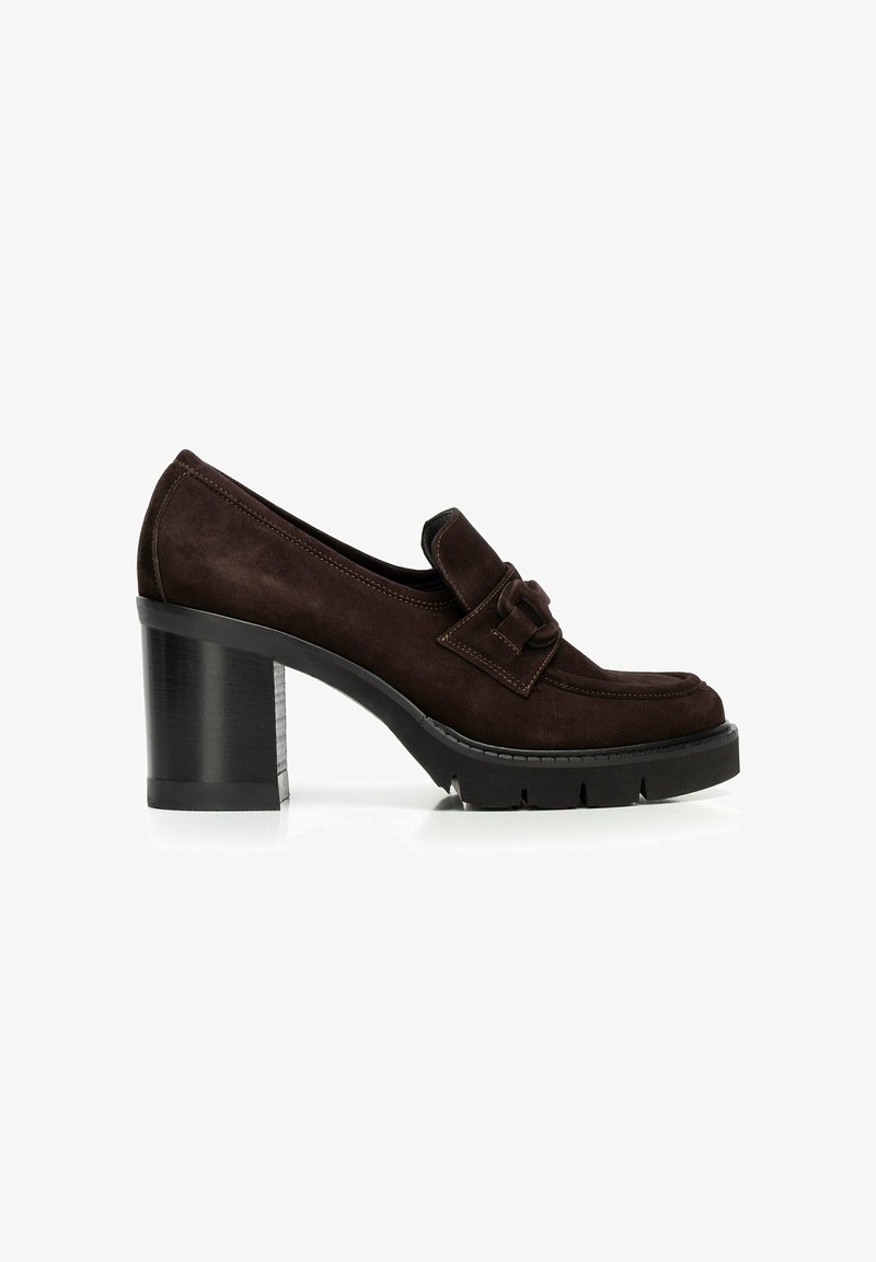 Mocassin en suède brun foncé avec un talon bloc épais, semelle en caoutchouc noir et détail de boucle décorative sur le dessus, présenté sur fond blanc.