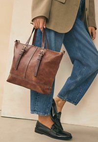 Brun læder tote bag med to håndtag, guldfarvet metal hardware, kontrasterende syninger, glat tekstur og en struktureret, rektangulær form.